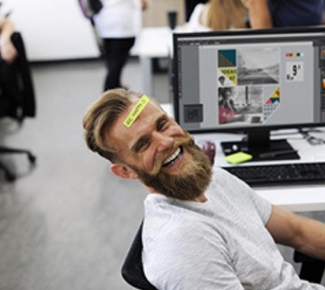 white bro dude with a big beard positioned in front of a computer, in a white tee leaning back and smiling