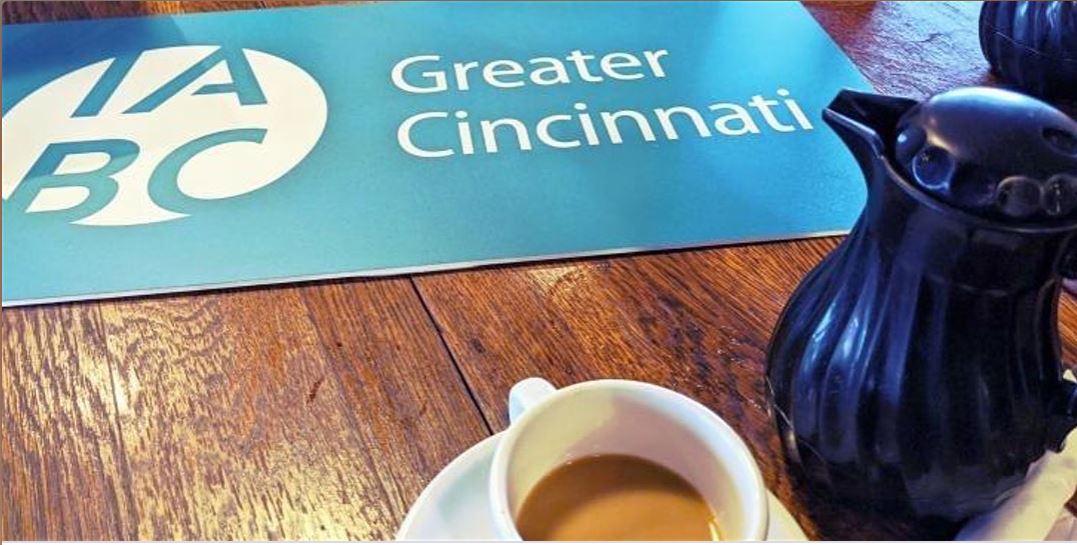 Wooden table with flat blue sign with IABC Greater Cincinnati logo, cup of coffee, and coffee pot